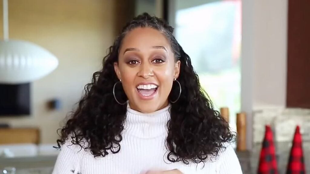 Tia Mowry in a white shirt with curly hair smiling into a camera.