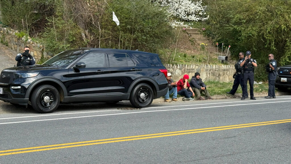 Multiple people detained by the Pentagon police outside REI Bailey's Crossing in Virginia sit on the ground while multiple officers film the bystander taking their picture