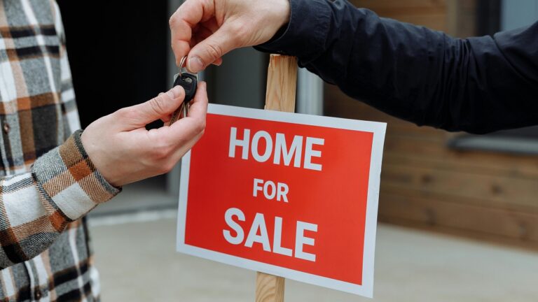 a person handing a key off, with a home for sale sign in the background
